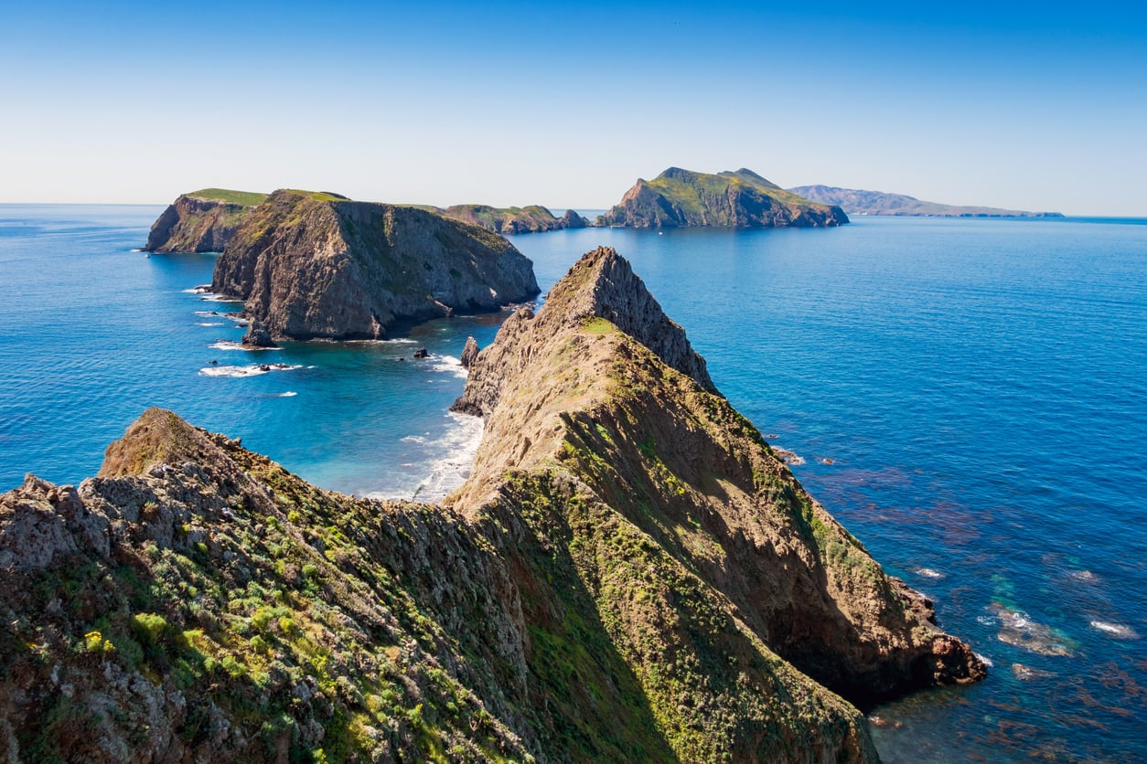 A rugged, rocky island with steep cliffs and green patches stretches into the blue ocean under a clear sky, with more islands visible in the distance—making it one of the most captivating travel destinations on the West Coast.
