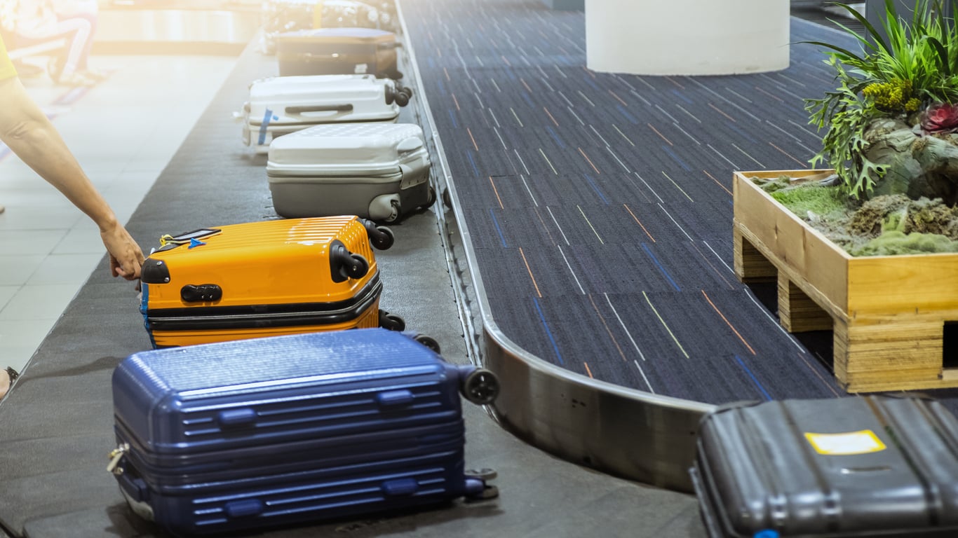A person reaches for a yellow suitcase on an airport baggage carousel, surrounded by other luggage in various colors—a vivid reminder of vacation costs waiting to be claimed alongside cherished memories.