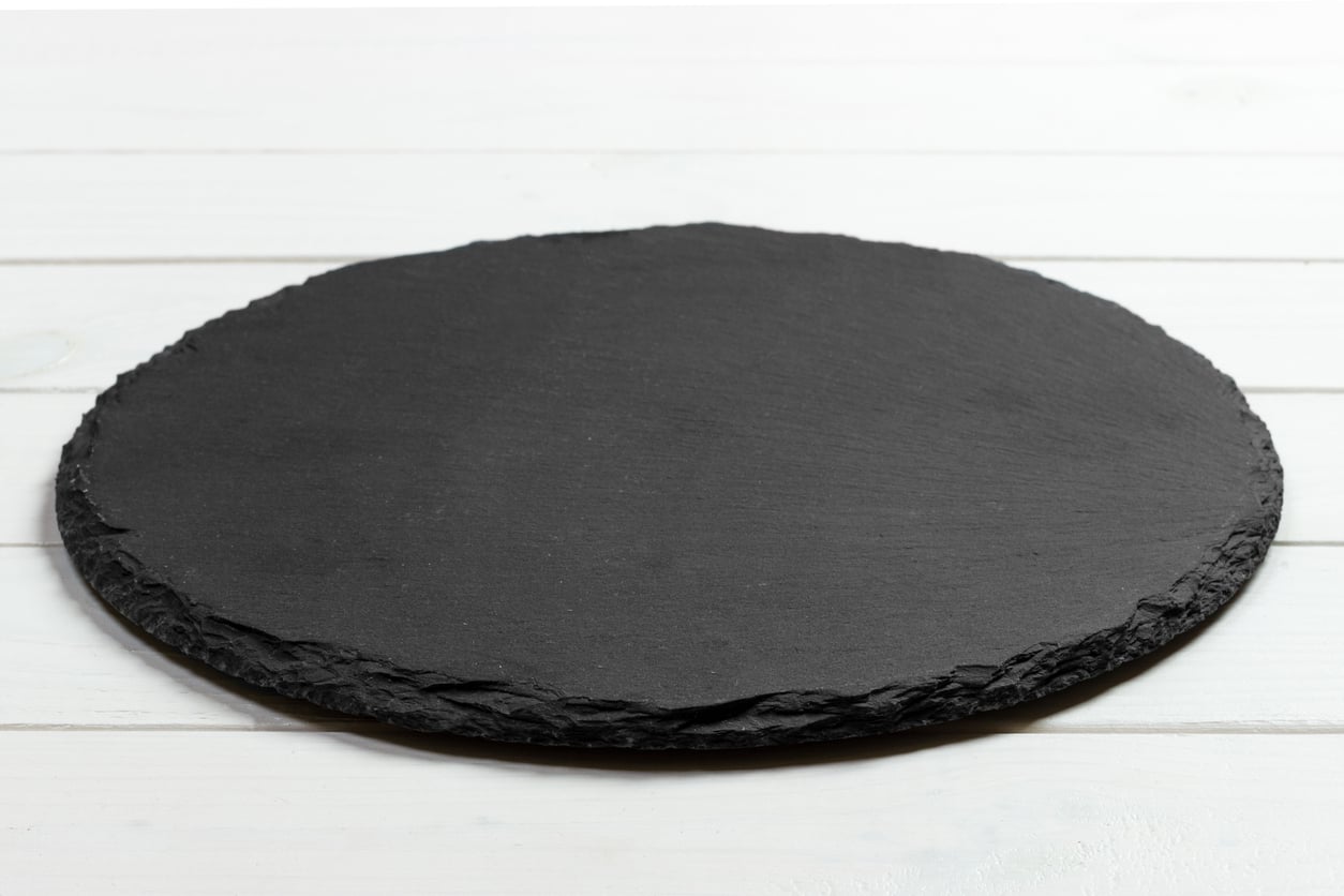 A round, flat, black slate serving board with a rough edge sits on a white wooden surface, adding a touch of elegance to your collection of kitchen items.