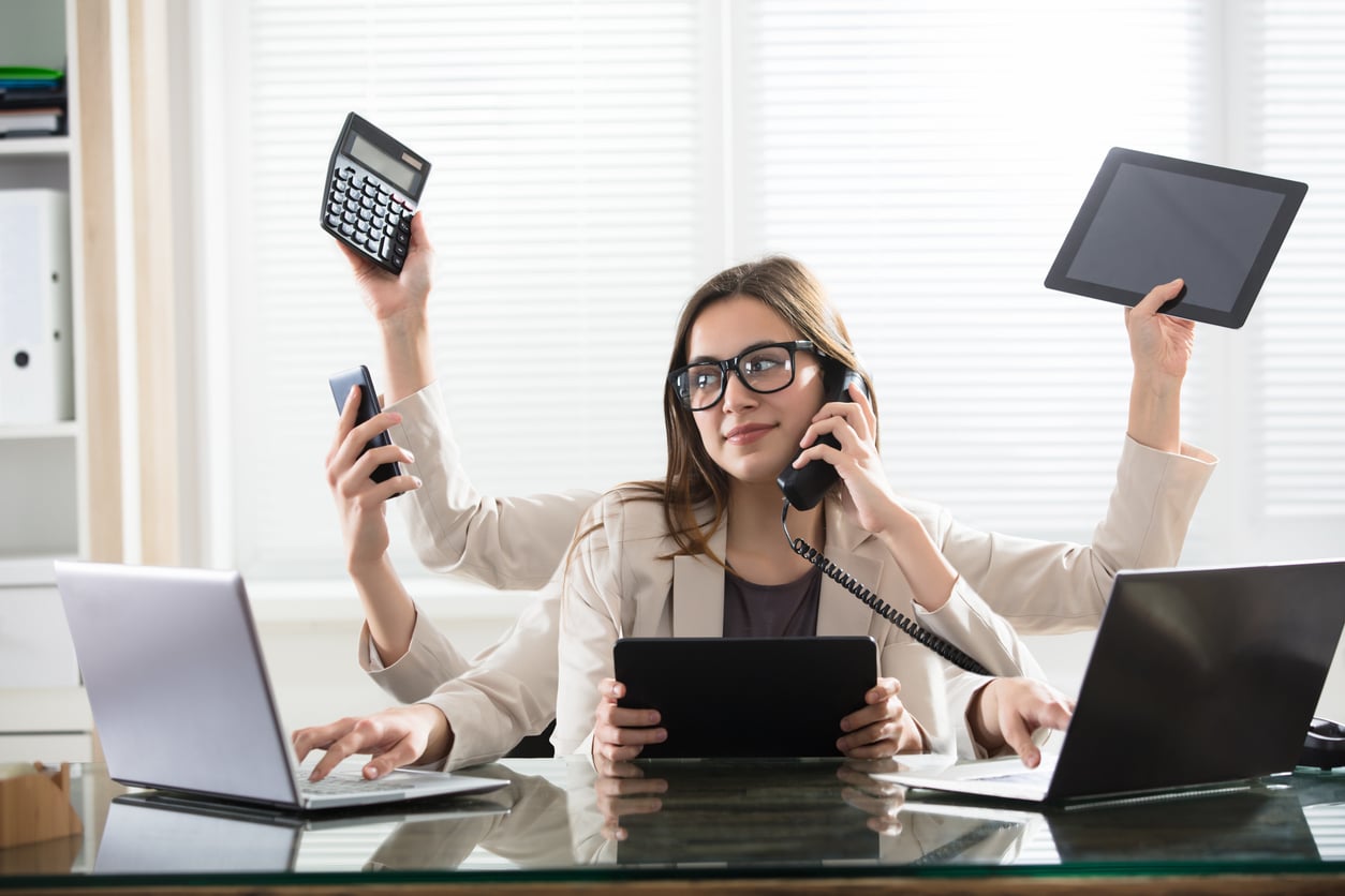 A woman with multiple arms sits at a desk, multitasking with a phone, tablet, calculator, laptop, and smartphone, embodying Baby Boomer beliefs about dedication and productivity in a busy work environment.