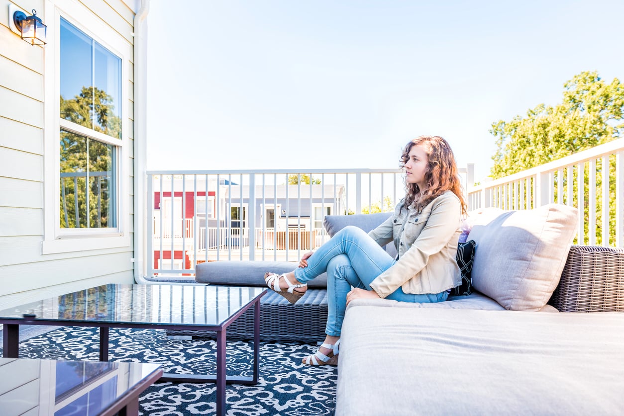 A woman sits alone on an outdoor sofa on a sunny balcony, facing a glass table. She looks thoughtful, surrounded by greenery and neighboring buildings in the background—a serene setting that showcases stylish outdoor home products.