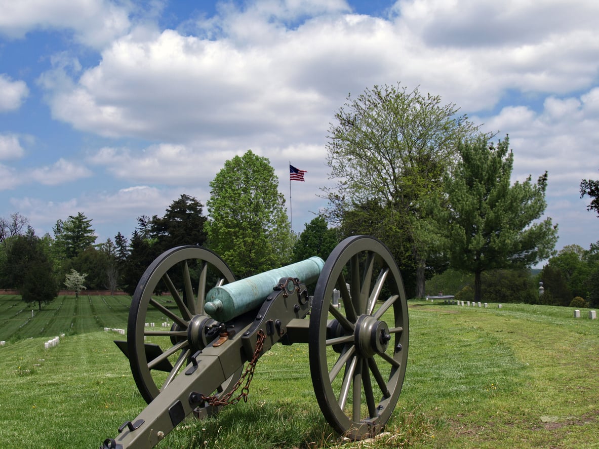 An old cannon sits on a grassy East Coast hill with gravestones in the background. An American flag waves on a tall pole among trees under a partly cloudy sky.