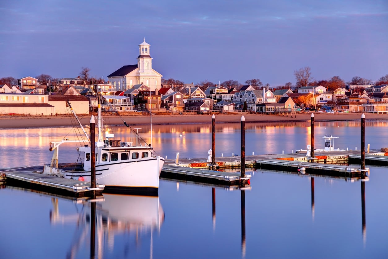 A white fishing boat is docked in a calm marina at sunset, with a row of houses and a large white church with a steeple in the background reflecting on the water, making this scene one of the most picturesque travel destinations for visitors seeking tranquility.