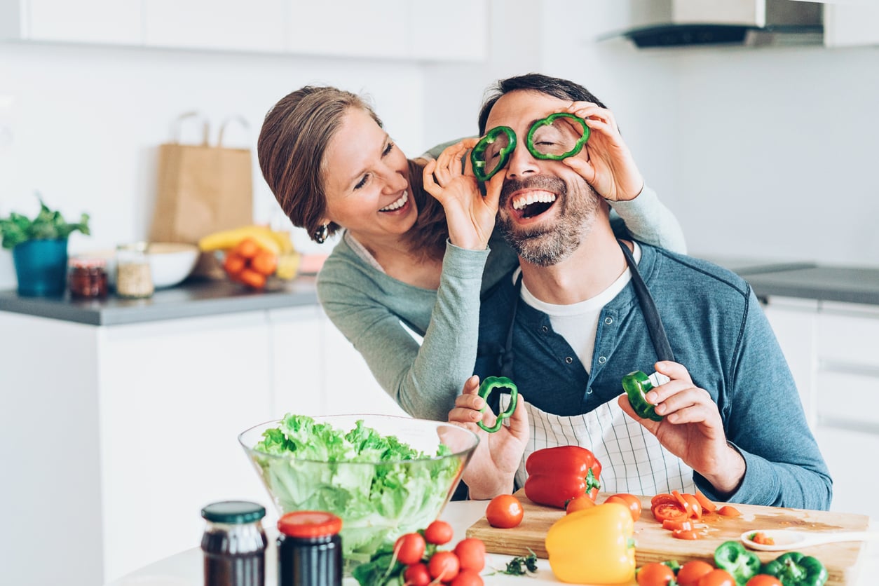 A smiling couple in a kitchen, with the man playfully holding green bell pepper slices over his eyes while the woman laughs behind him. Fresh vegetables and salad are spread on the counter in front of them.