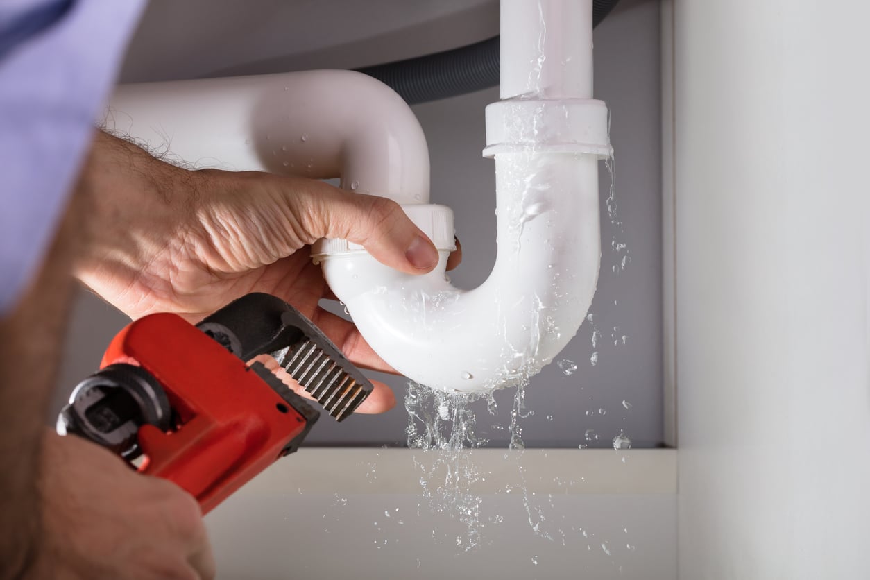 A person uses a red pipe wrench to fix a leaking white plastic pipe under a sink, with water spraying out from the joint.