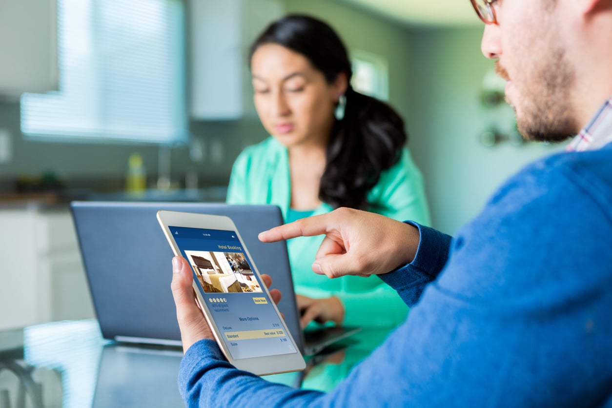 A man uses a tablet to browse home listings and avoid common hotel booking mistakes, while a woman works on a laptop in the background. They are seated at a table in a bright, modern room.