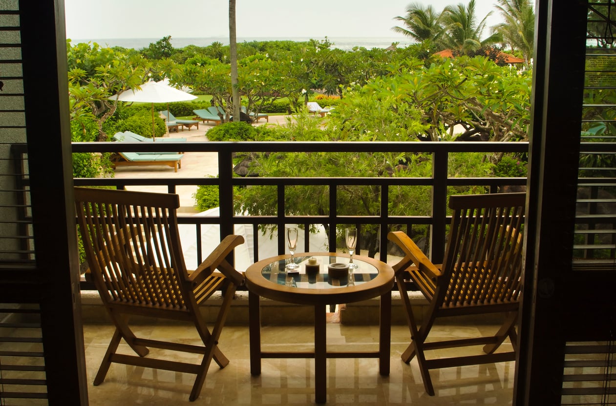 Two wooden chairs and a table with glasses on a balcony overlook a lush garden with lounge chairs, umbrellas, and palm trees. The ocean is visible in the distance beyond the greenery.