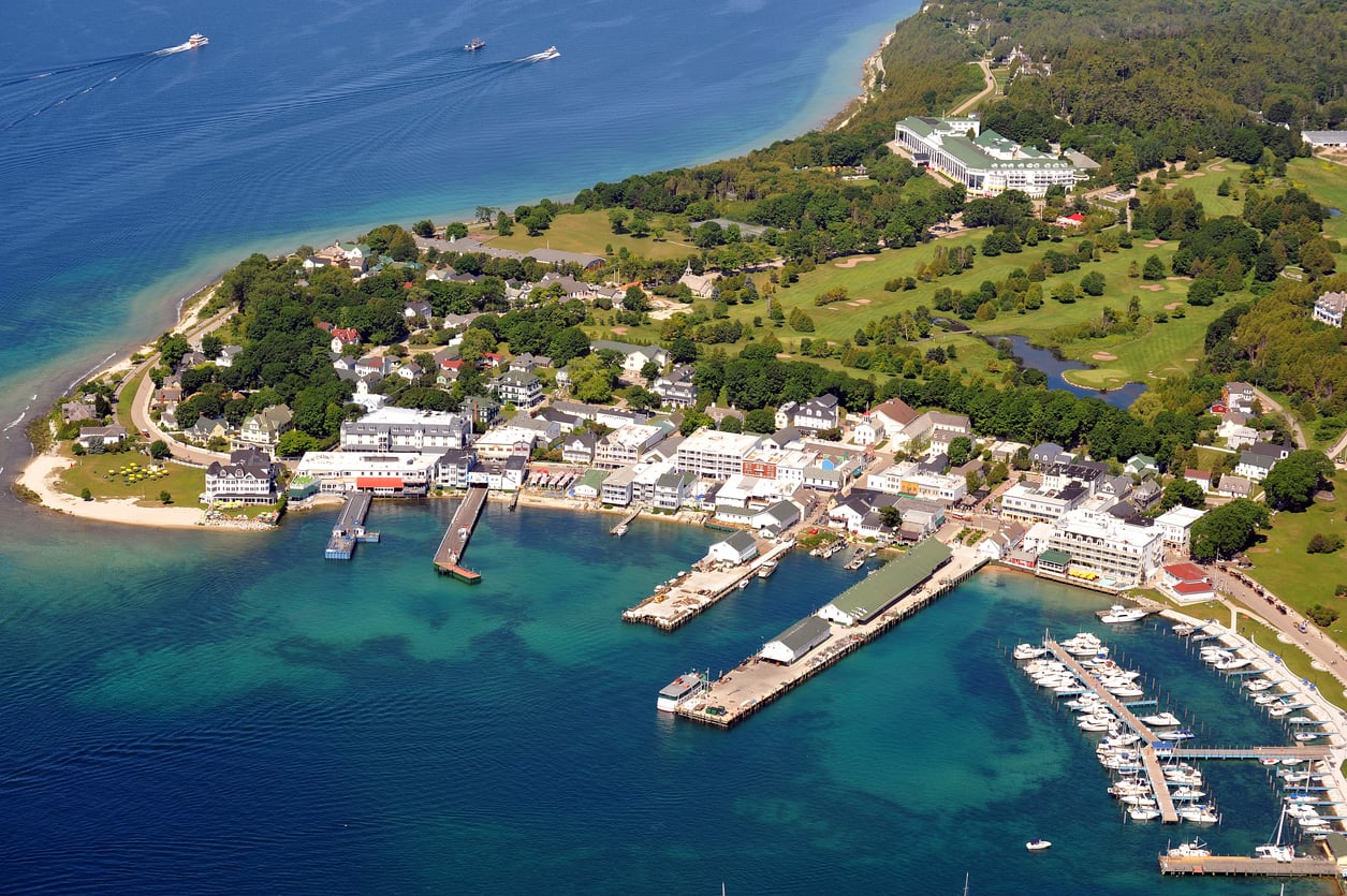 Aerial view of a small island town with docks, marinas, white buildings, green trees, and clear blue water. Boats are visible in the harbor. A large hotel or resort is on a hill in the background.