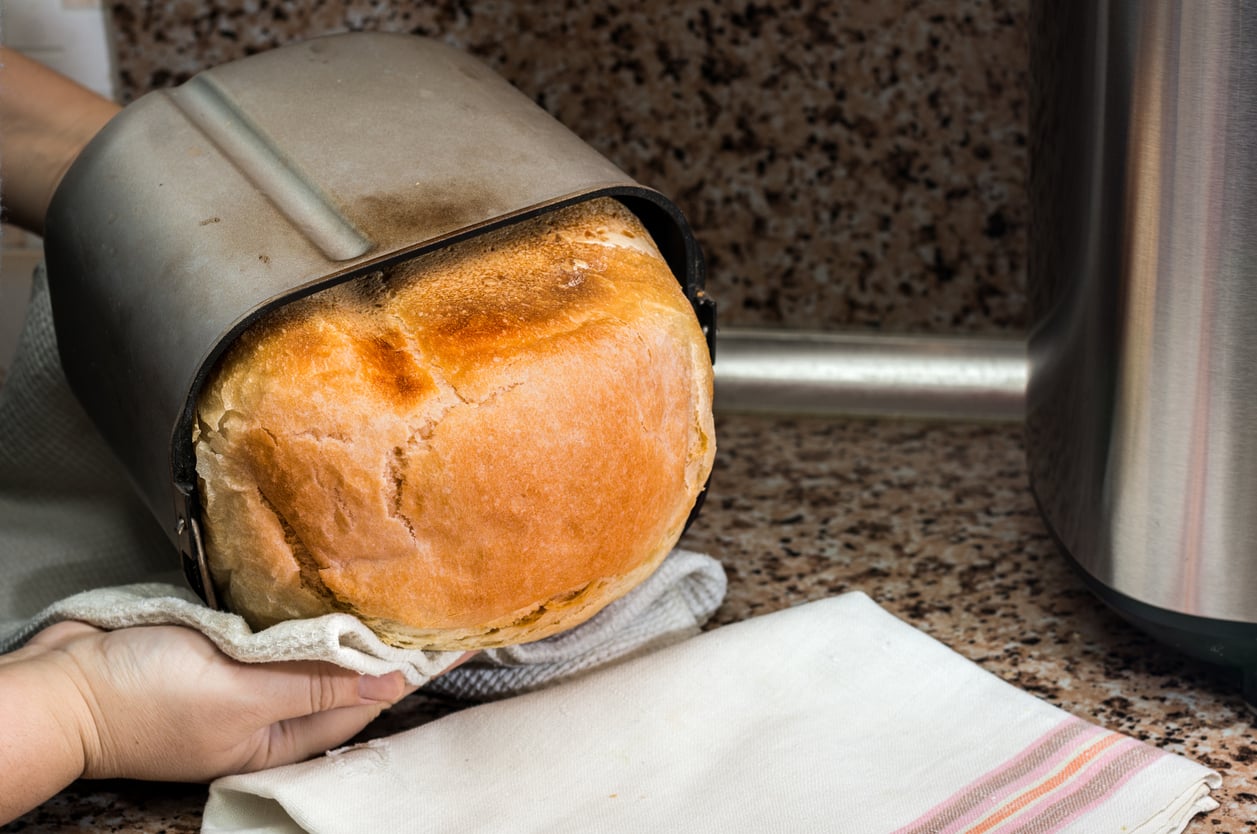 A person removes a freshly baked loaf of bread from a bread machine pan onto a kitchen counter topped with a striped towel, surrounded by various kitchen items, with a brown granite backsplash in the background.