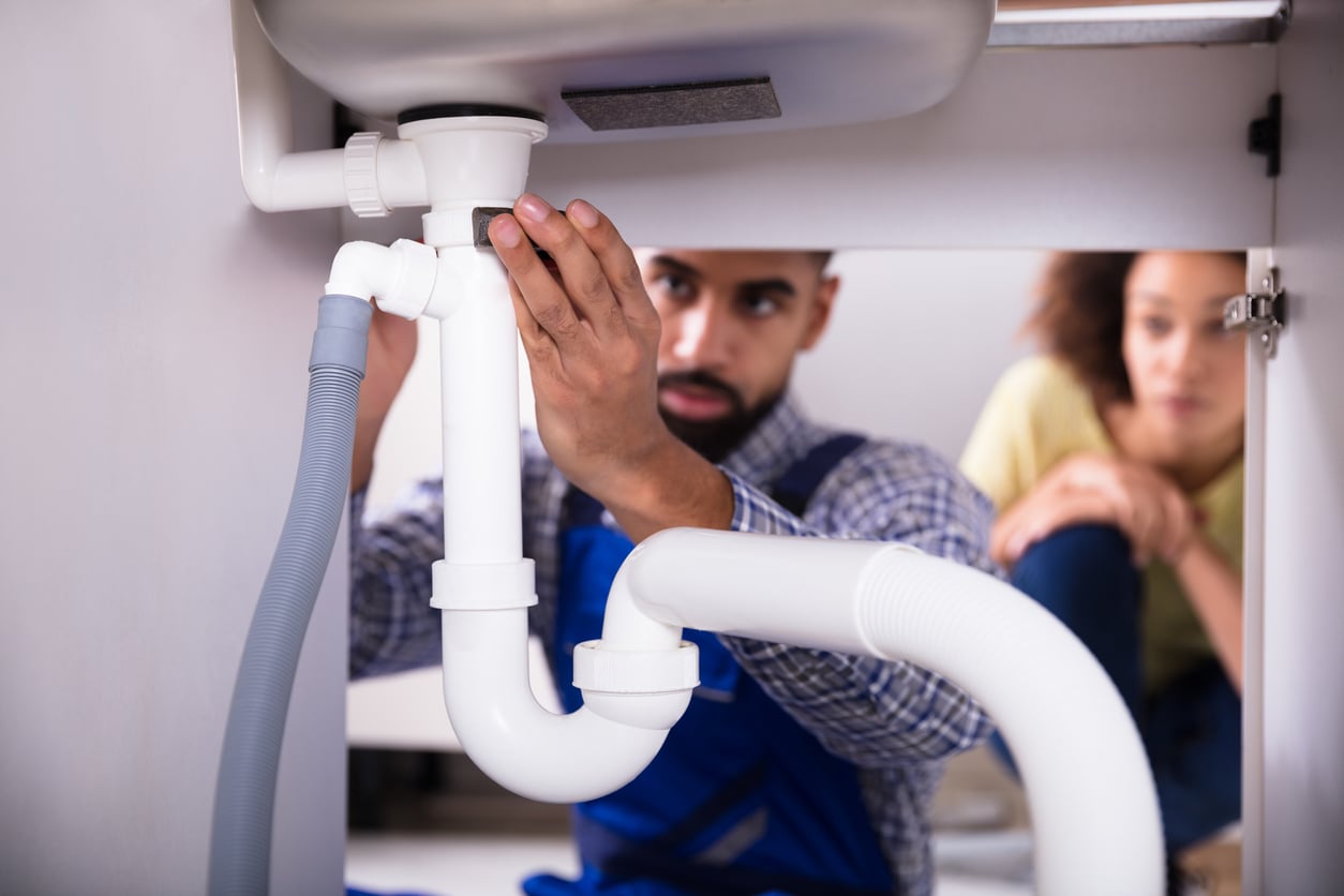 A plumber in blue overalls repairs white pipes under a sink, while a woman watches attentively in the background.