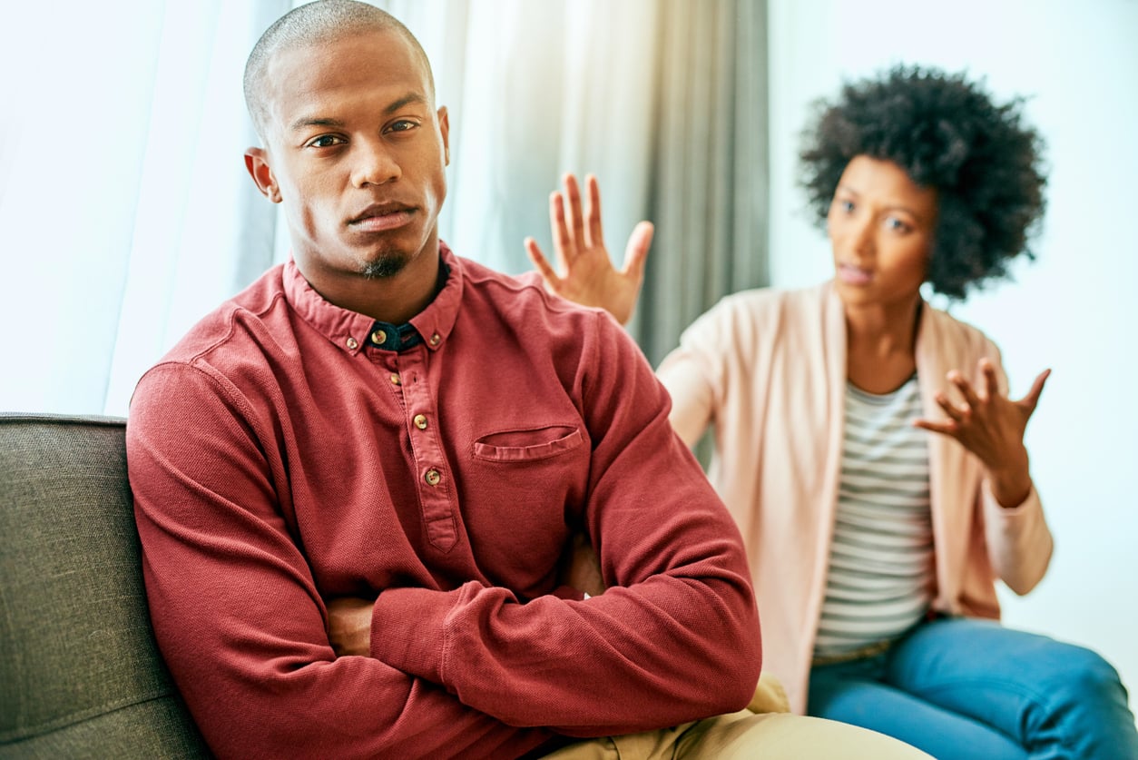 A man sits with arms crossed, looking serious—perhaps reflecting on money saving habits—while a woman in the background gestures with her hands as if explaining or arguing.