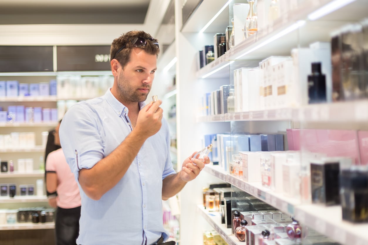 A man in a light blue shirt, known for his money saving habits, smells a perfume tester bottle while shopping in a store aisle filled with various perfume boxes and bottles.