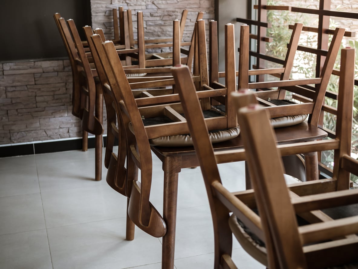 Wooden chairs are stacked upside down on top of wooden tables inside a room with tiled floors, stone-patterned walls, and sunlight coming through large windows with wooden bars. Restaurant chain closures.