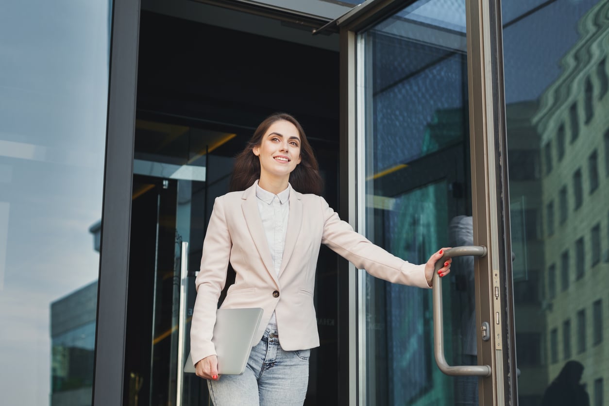 A woman in a light blazer and jeans, holding a laptop, smiles as she exits a modern glass building, holding the door open—capturing a moment that feels reminiscent of life before smartphones, when face-to-face interactions and simple gestures connected people most.
