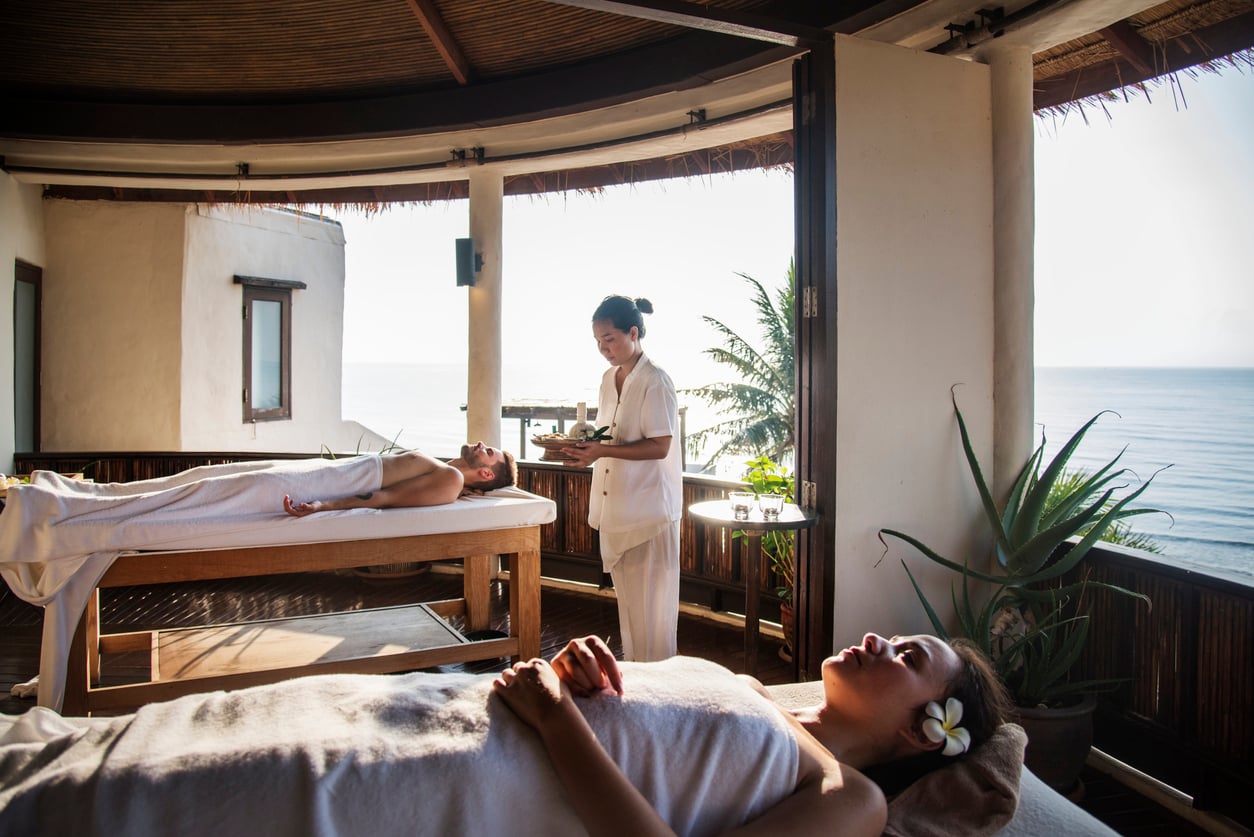 Two people relax on massage tables in an open-air spa room overlooking the ocean, grateful to have avoided hotel booking mistakes, while a masseuse prepares treatment in the serene, sunlit space.