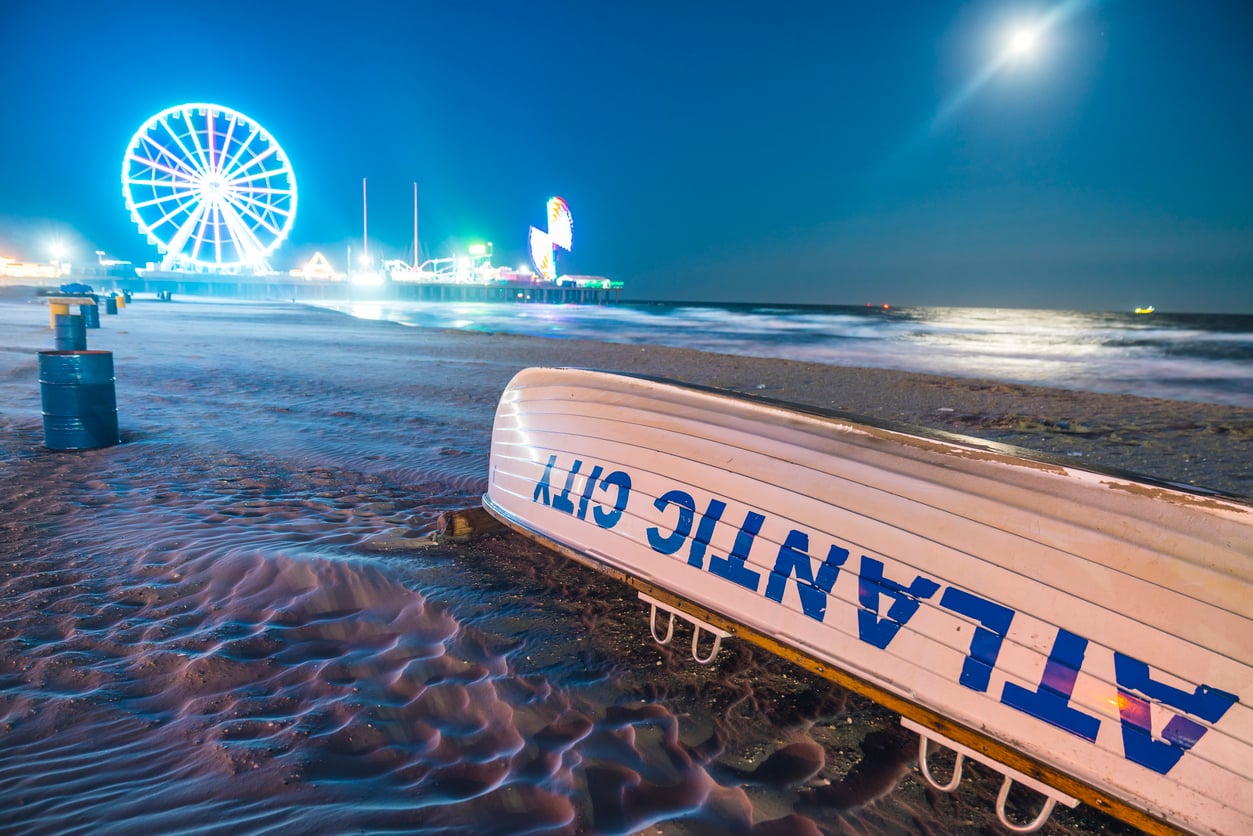 A lifeboat labeled "Atlantic City" sits on the sandy beach at night, with bright amusement park lights and a Ferris wheel glowing in the background under a moonlit sky—capturing the vibrant allure of one of America's most iconic and affordable travel destinations.