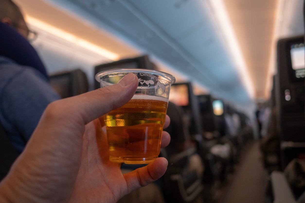 A hand holding a plastic cup filled with beer inside an airplane cabin, with rows of seats and passengers visible in the background.