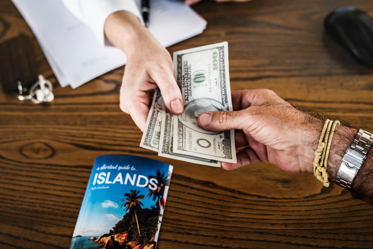 A person hands cash to another person across a wooden table, perhaps settling vacation costs, with a travel guide titled "Islands" and some papers and a pen in the background.