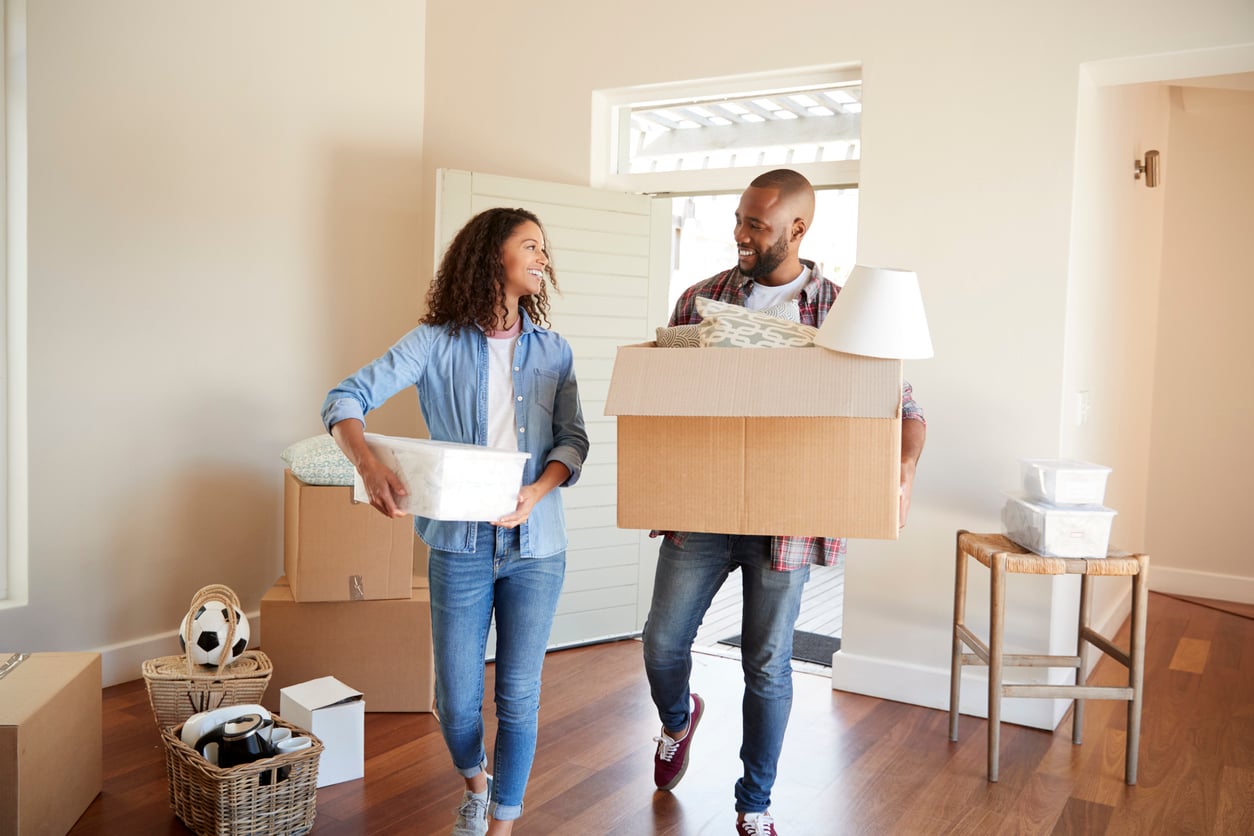 A smiling couple carrying boxes and household items into a bright, unfurnished room, suggesting the benefits of renting as they move into a new home. Other packed boxes and belongings are scattered on the floor around them.