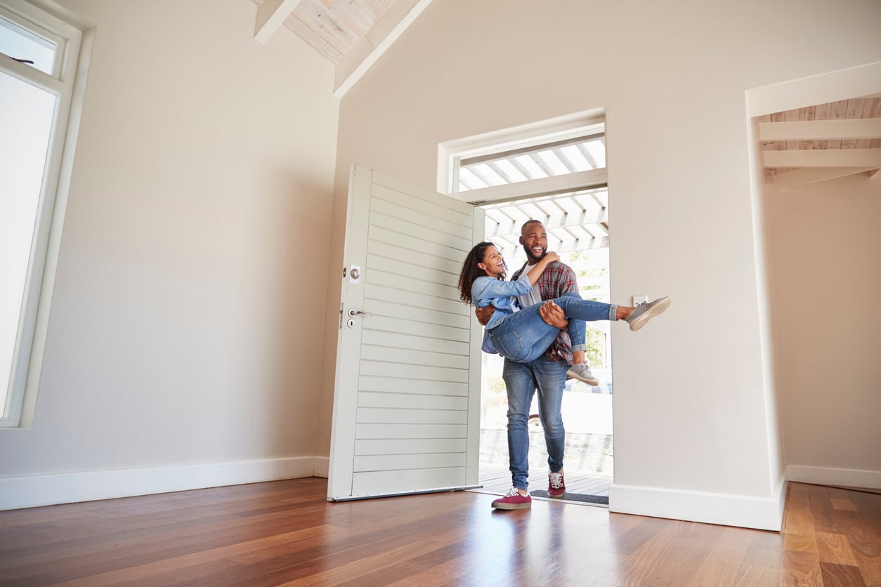 A man carries a smiling woman across the threshold of a bright, empty room with wooden floors, capturing the excitement and benefits of renting as they move into their new home together.