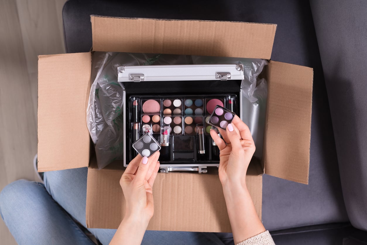A person opens a cardboard box containing a makeup kit, holding two small eyeshadow palettes above the kit, which includes various cosmetics like lipsticks, blush, and brushes.
