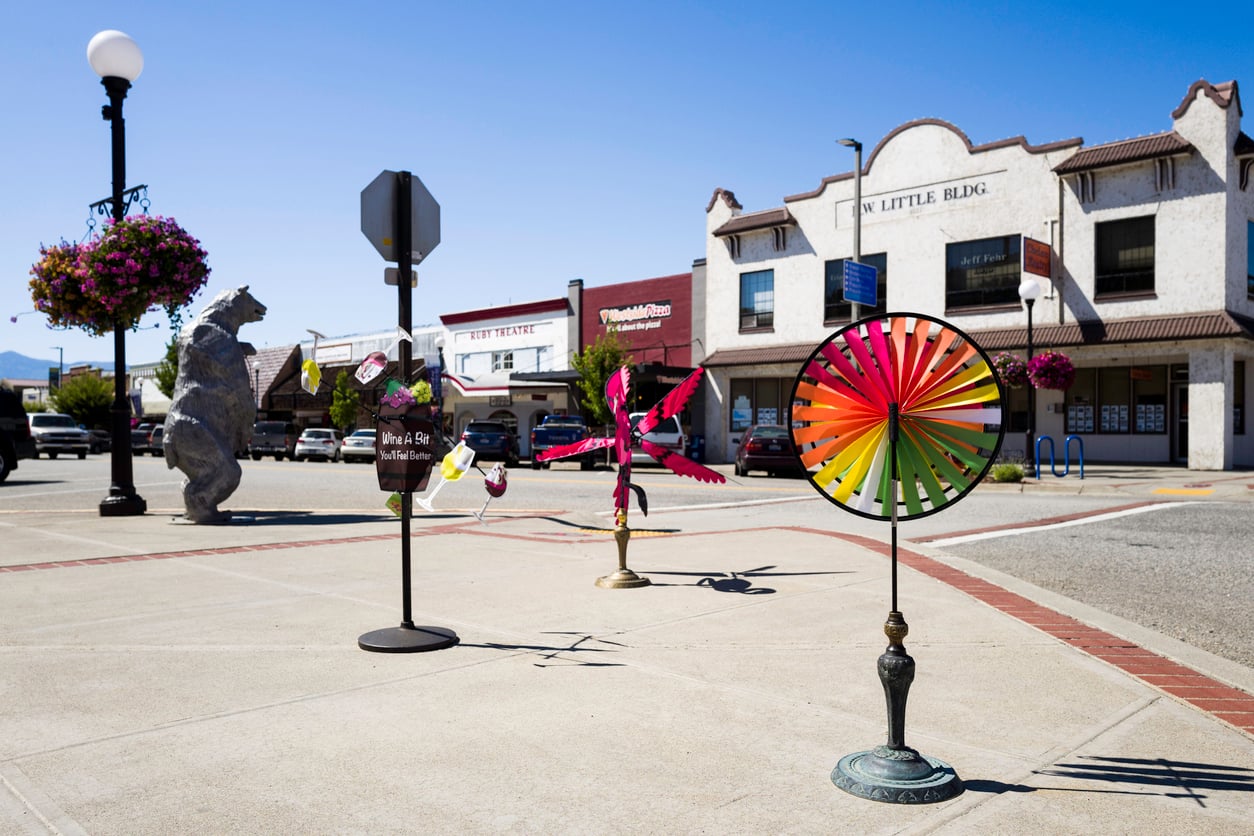 Colorful pinwheels and sculptures are displayed on a sunny downtown sidewalk with hanging flower baskets, parked cars, and historic buildings in the background.