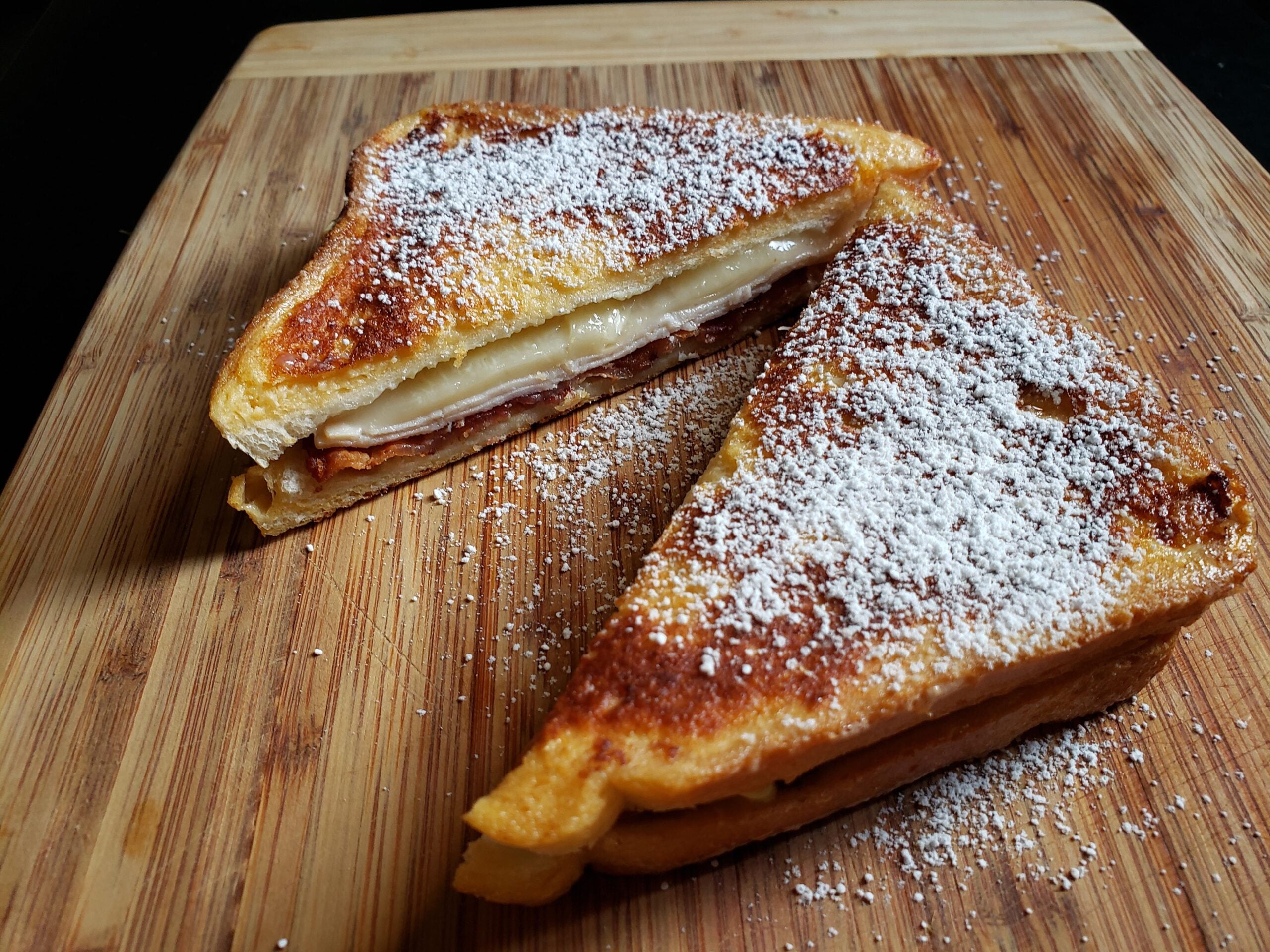 A grilled sandwich with melted cheese and ham, cut in half and dusted with powdered sugar, sits on a wooden cutting board.