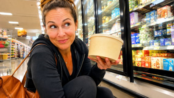 Woman crouching in a grocery store, smiling and holding a large beige container in front of a refrigerated section filled with various dairy products&mdash;she knows the smart shopper&rsquo;s secret: there are foods to never buy at full price.