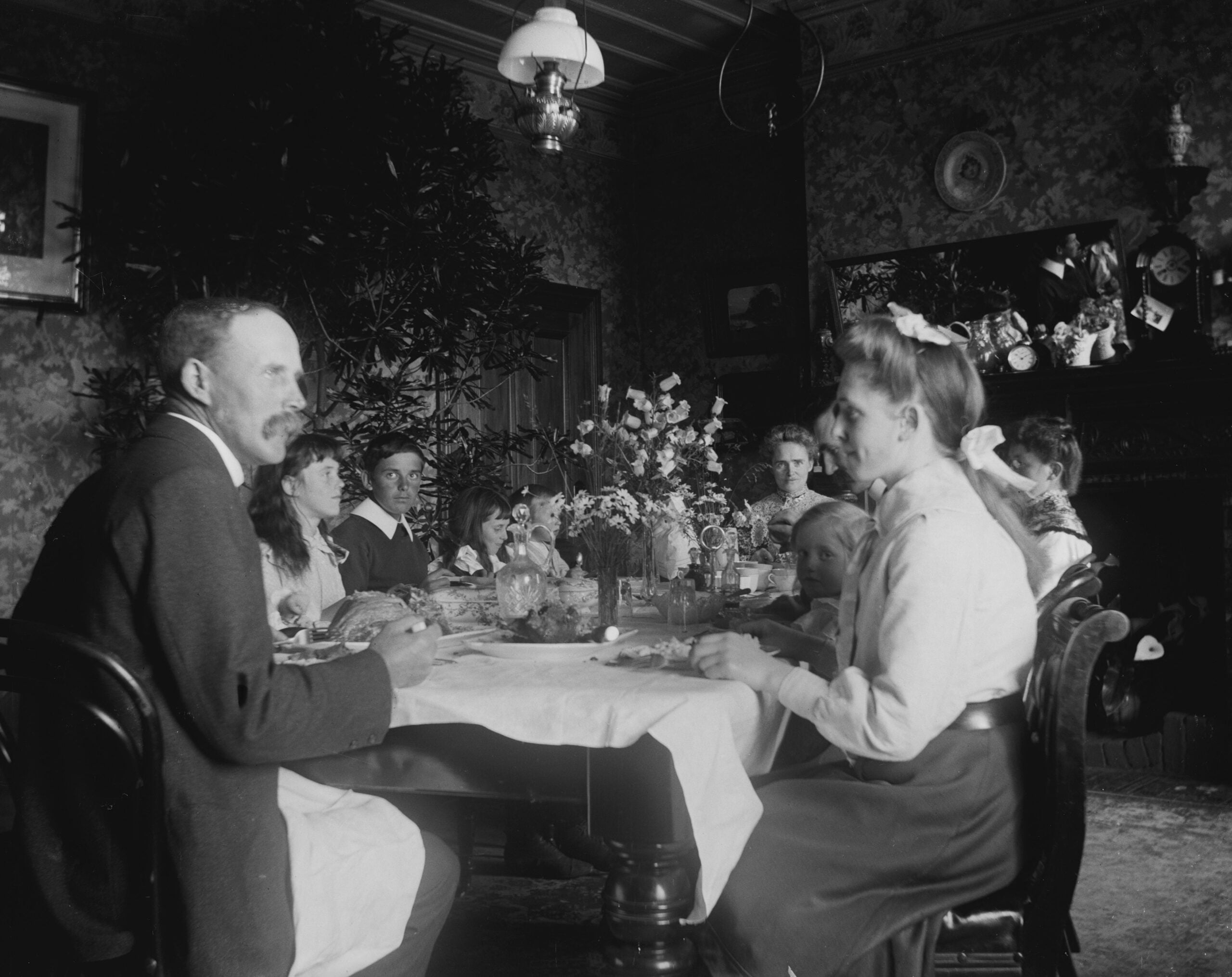 A black and white photo of a family gathered around a dining table set for a meal in a richly decorated room, evoking nostalgic dinners, with flowers in the center and framed pictures on the walls.