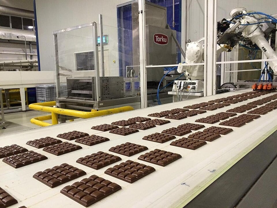 Rows of chocolate bars move along a conveyor belt in a factory, with robotic machinery and equipment visible in the background.