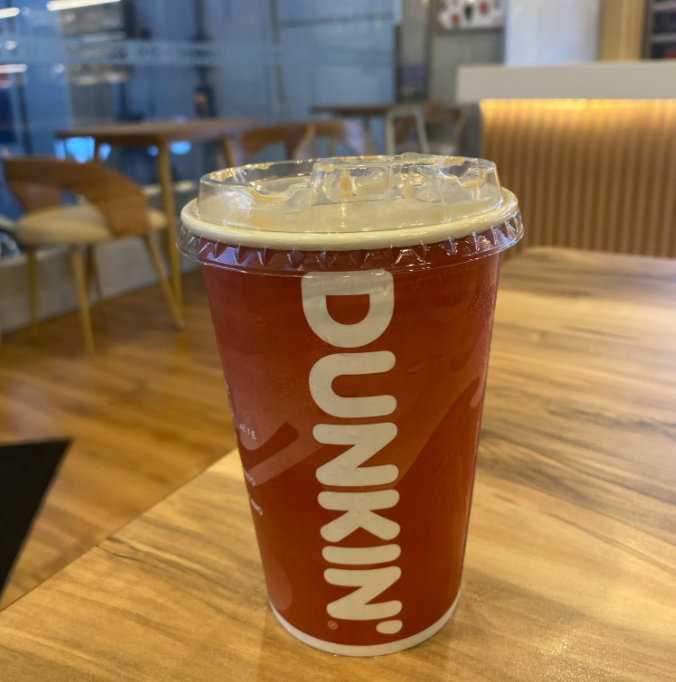 A red Dunkin' cup with a plastic lid sits on a wooden table in a cafe, capturing the cozy vibe often found in coffee houses, with chairs and tables visible in the blurred background.