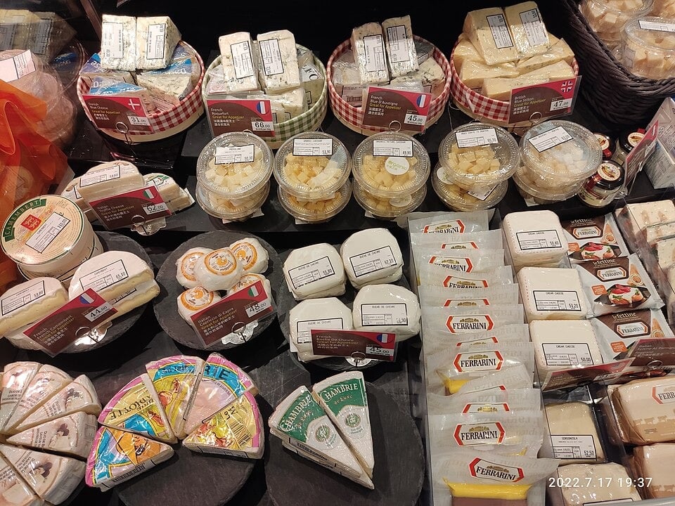 A supermarket display featuring assorted cheeses, including wedges, wheels, and packaged blocks, with visible labels and various countries' flags, neatly arranged on black shelves.