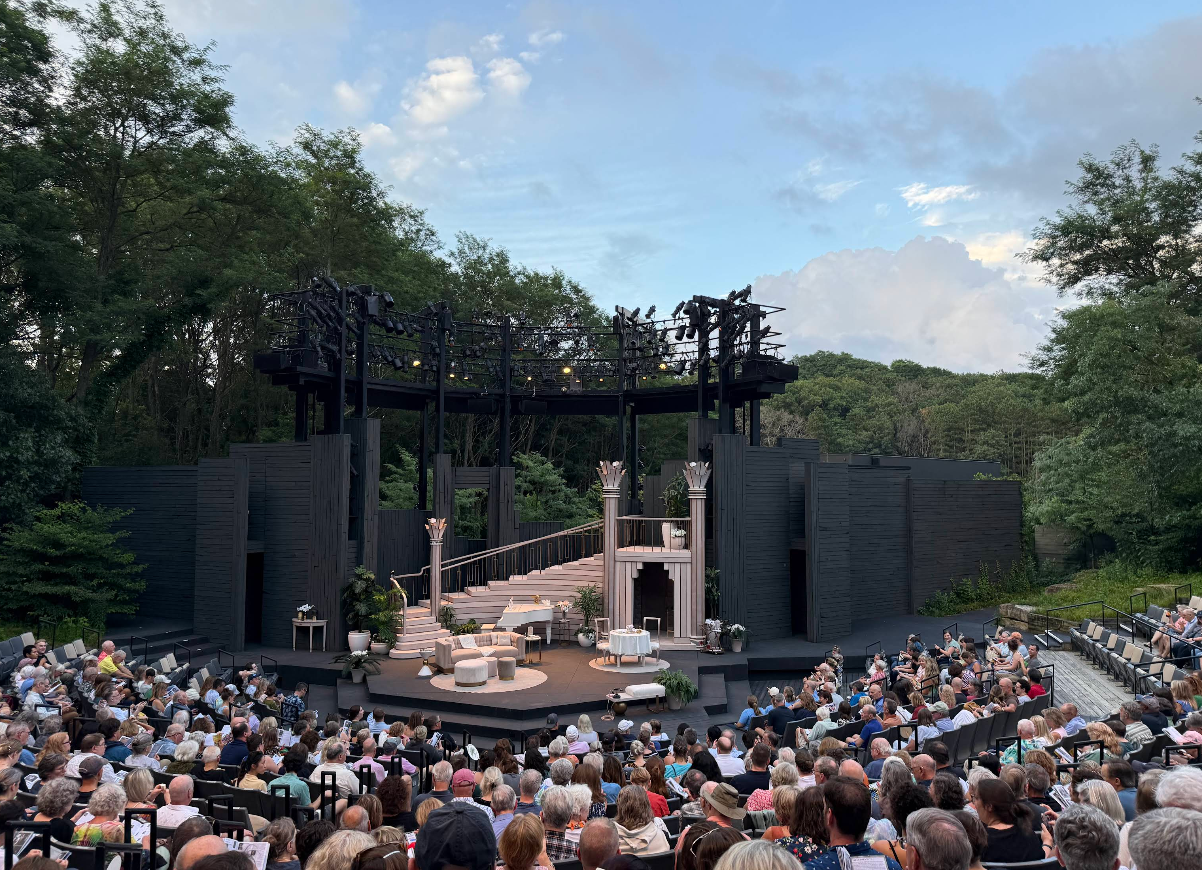 An outdoor live theater hosts a large audience before a modern stage set with tall columns, a spiral staircase, and greenery, all nestled among dense trees beneath a partly cloudy sky.