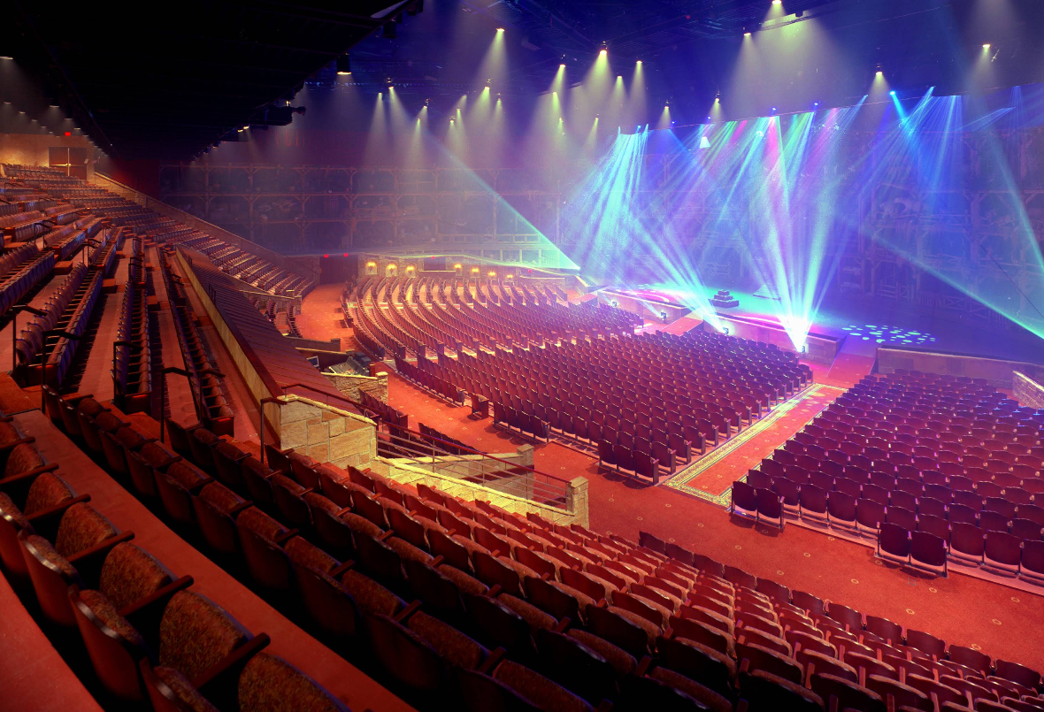 A large, empty live theater with rows of red seats, dramatic stage lighting casting colorful beams, and an illuminated stage, all viewed from an upper seating area beneath a high ceiling.