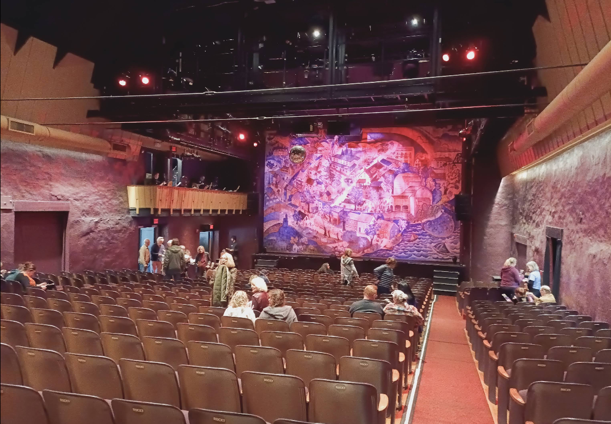 People are seated and standing in an auditorium with rows of brown chairs facing a colorful curtain on stage, ready for live theater. The purple walls and balcony on the left add to the ambiance, while red lights illuminate the vibrant scene.