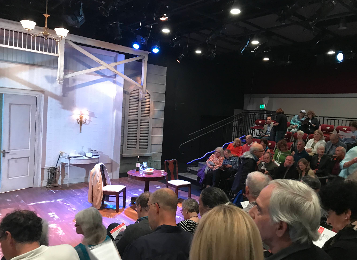 An audience sits in a theater with red seats, eagerly awaiting the start of live theater. The stage is set with a small table, two chairs (one draped with a coat), and a wall featuring a door, shutters, and a hanging light fixture.