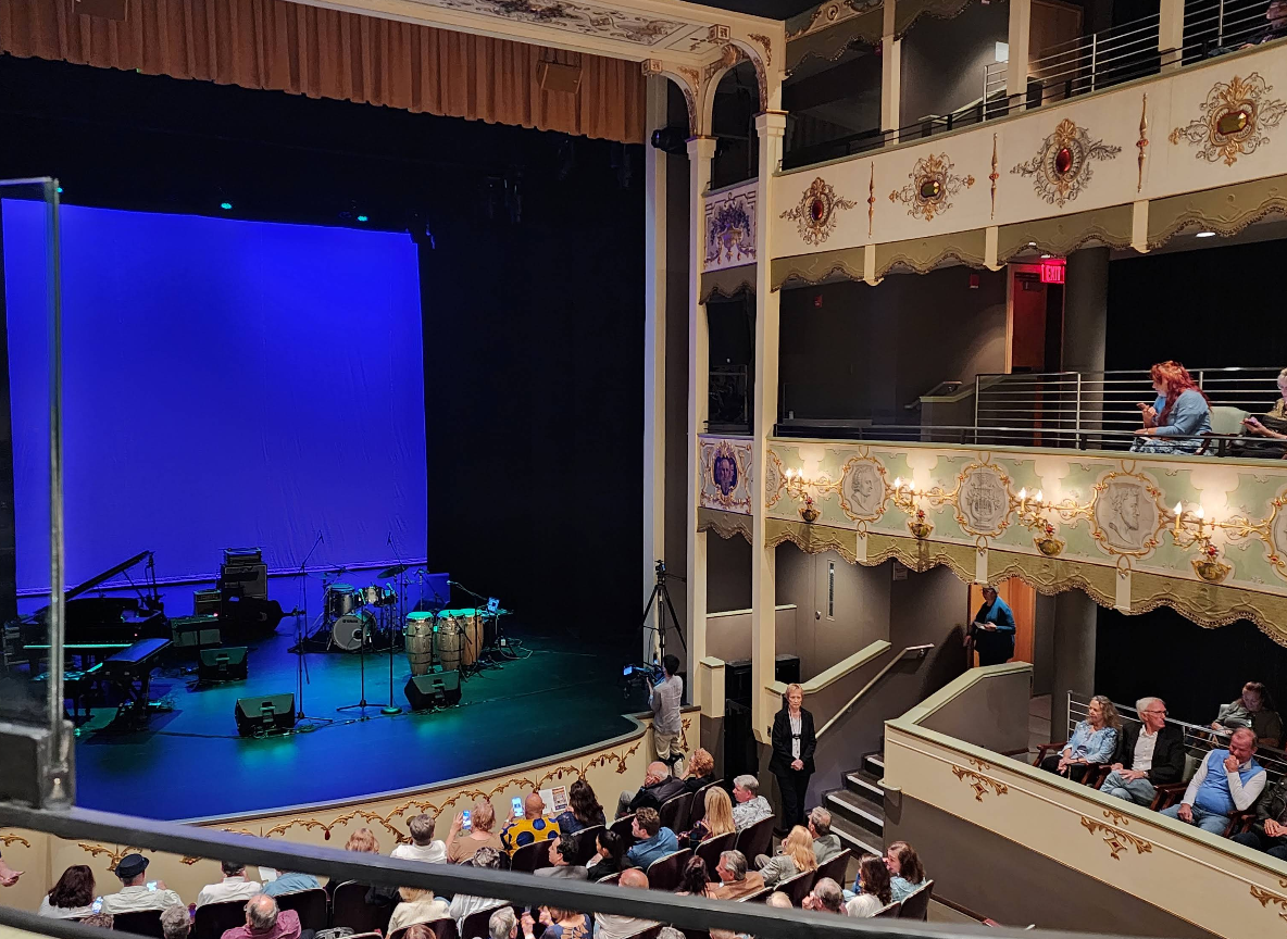 A live theater with ornate balconies and a seated audience faces a stage lit with blue lighting. Musical instruments, including drums, keyboard, and guitar, are set up on stage, awaiting a performance.