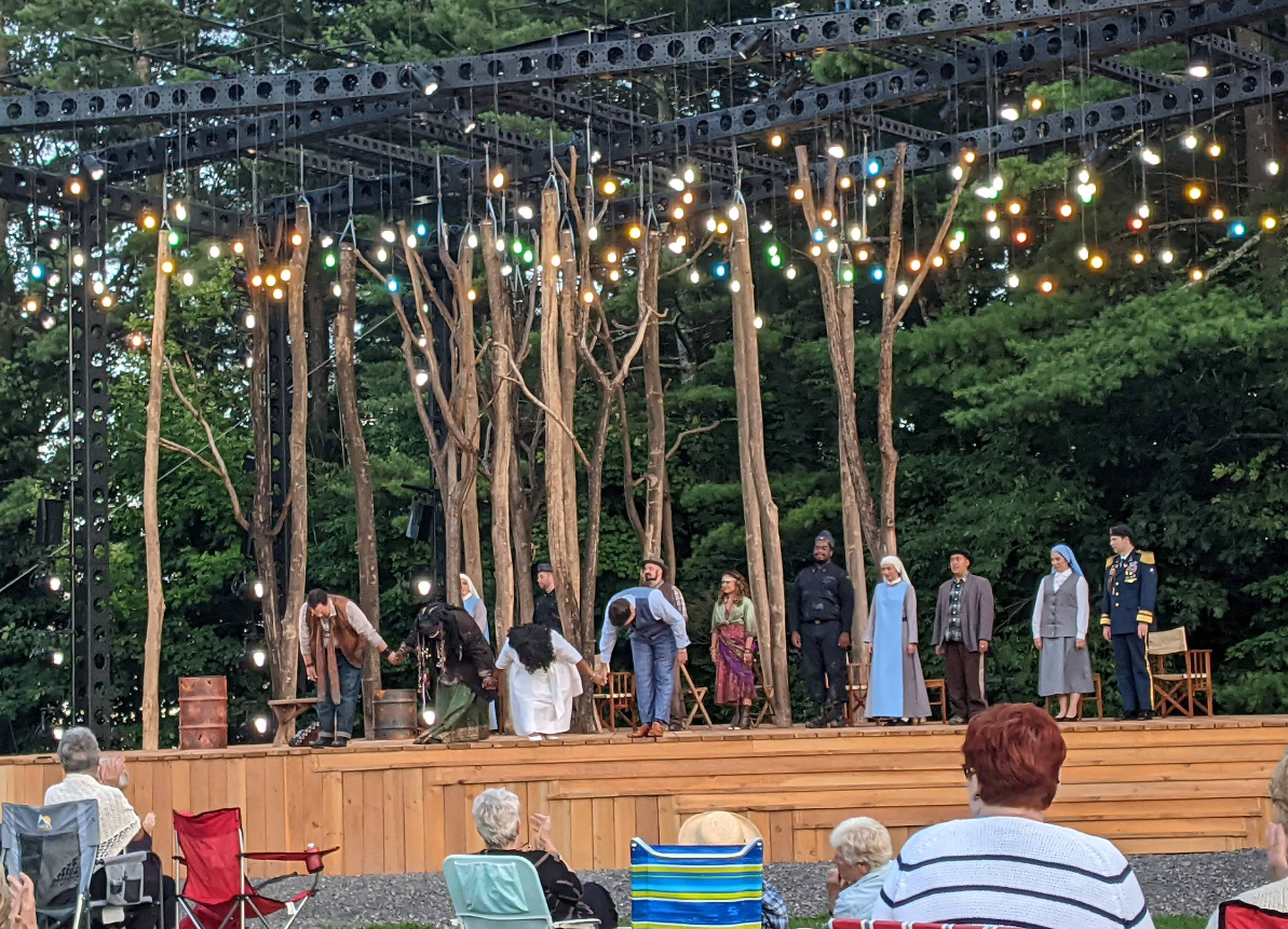 Actors in costume perform live theater on an outdoor stage decorated with tree trunks and colorful hanging lights, while an audience watches from folding chairs. The stage is set against a backdrop of dense green trees.