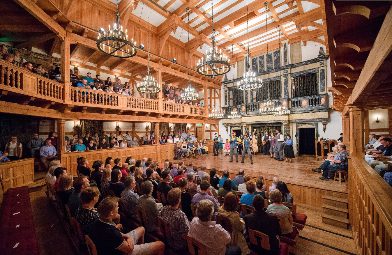 Audience watches a captivating Live Theater performance on a wooden stage inside a theater with balconies, chandeliers, and a high ceiling. The venue is full, with people seated all around and actors performing in the center.