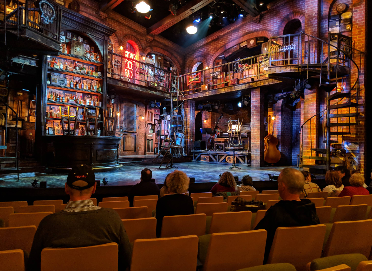 View from the audience of a cozy, warmly lit Live Theater set designed as a book-filled bar with spiral stairs, shelves, tables, neon signs, and musical instruments, as people wait for a performance to begin.