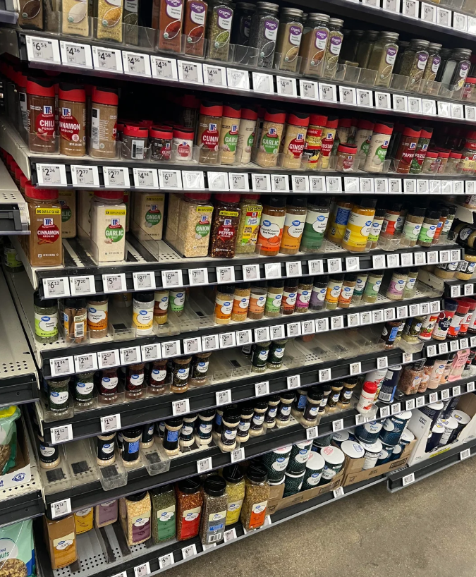A Walmart grocery store spice aisle displays various spices and seasonings in jars and containers arranged on shelves, each labeled with prices. The well-stocked shelves offer a wide variety of options.