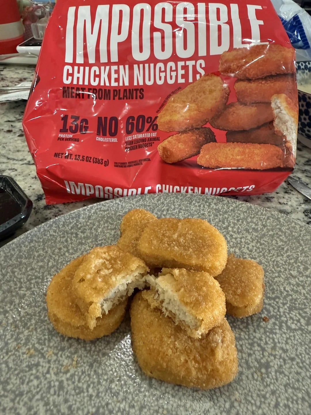 Four Impossible chicken nuggets, one broken in half, are on a gray plate. The red packaging, labeled “Impossible Chicken Nuggets,” is visible behind them on a kitchen counter.
