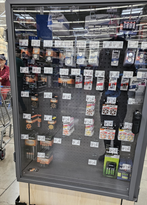 A locked display case at Walmart contains various brands and sizes of batteries, including Energizer and Duracell. Price tags are visible, some shelves are partially empty, and a shopper with a cart is in the background.