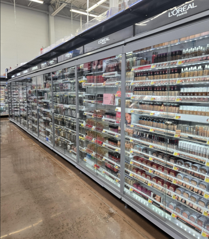 A Walmart supermarket aisle features glass display cases filled with various L'Oréal Paris cosmetics, including foundations, lipsticks, and powders, all neatly arranged on shelves behind closed doors.
