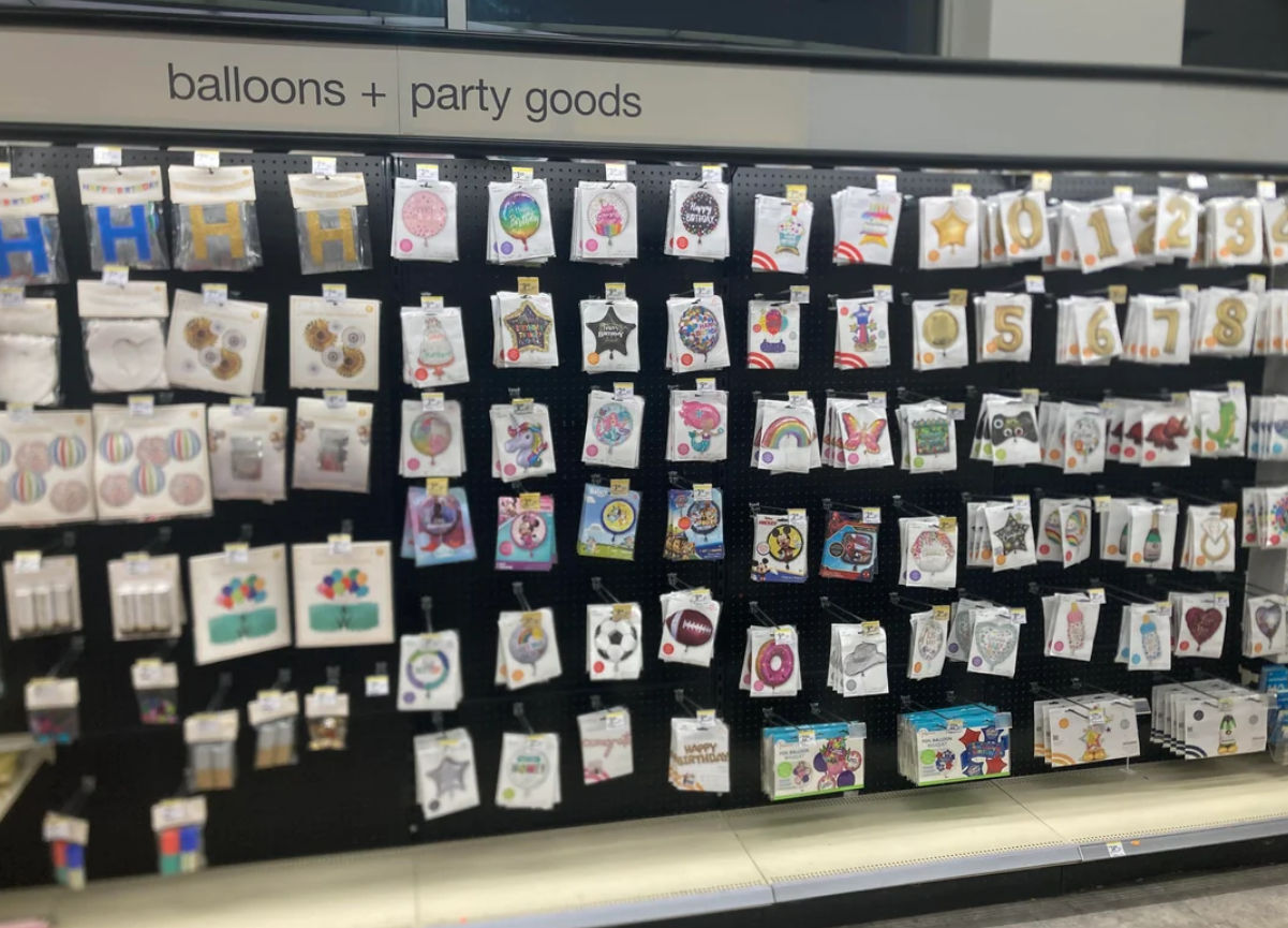 A Walmart store display labeled "balloons + party goods" shows packs of colorful balloons, number balloons, and various party decorations hanging on hooks against a black background, with shelves below holding more party items.
