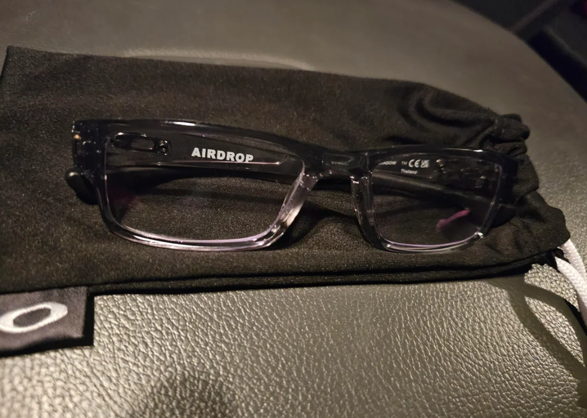 A pair of black rectangular Oakley eyeglasses from Walmart with "AIRDROP" printed on the inside of the left arm, resting on a black soft case on a textured gray surface.