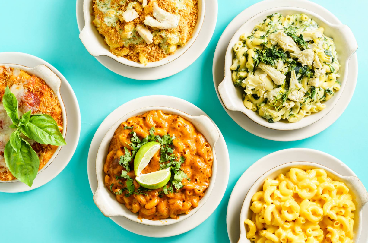 Five bowls of mac n cheese dishes with various toppings, including lime wedges, basil leaves, and greens, are arranged on a light blue background. Each bowl features a unique twist on classic macaroni and cheese.