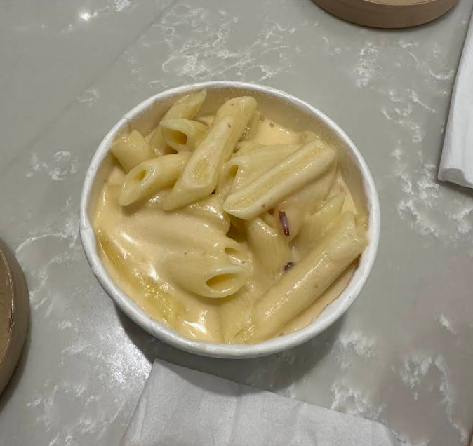 A white bowl filled with penne pasta in creamy mac n cheese sauce sits on a gray marble surface, next to a folded white napkin.