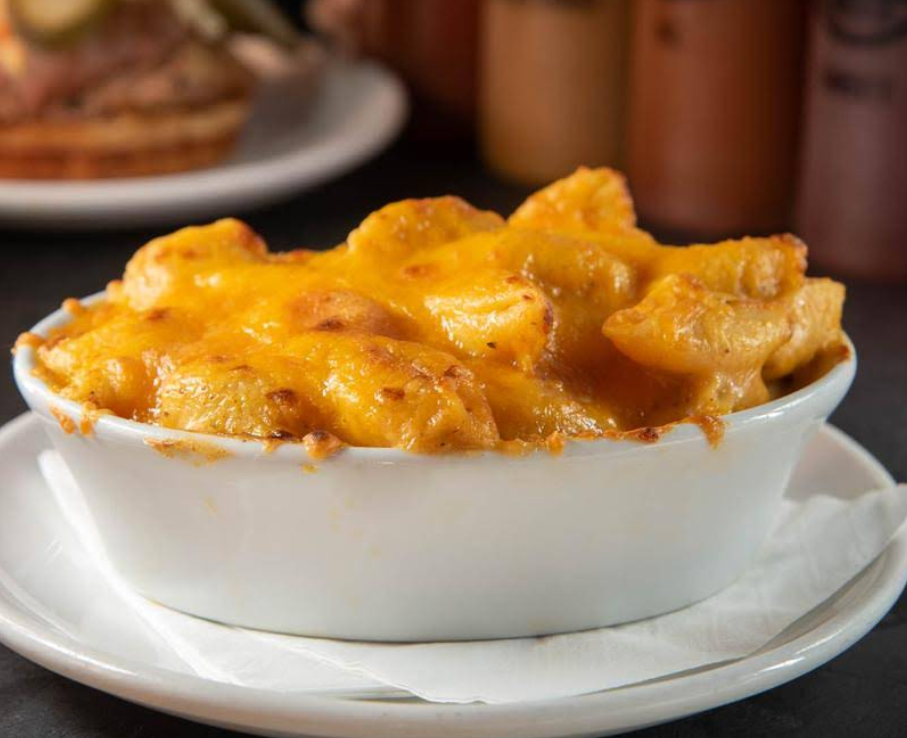 A white oval dish filled with creamy mac n cheese sits on a white plate with a napkin, topped with golden-brown melted cheese. Blurred bottles and food are visible in the background.