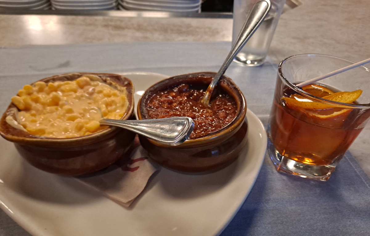 Two ceramic bowls—one filled with creamy mac n cheese, the other with hearty chili, each with a spoon. Beside them sits a cocktail glass garnished with an orange slice and cherry, all arranged on a countertop.